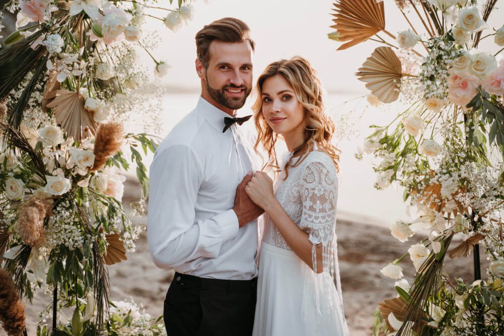 A couple dressed in wedding attire sharing a moment by a floral archway on the beach, photographed by their Destination Wedding Travel Agent.