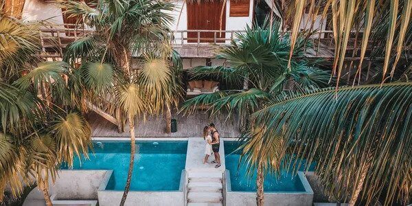 Two individuals on their honeymoon standing by a tranquil pool surrounded by lush palm trees in a tropical resort setting.