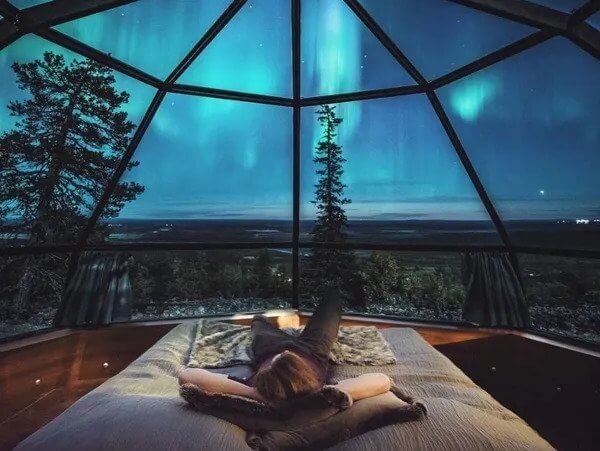 Person relaxing on a bed inside a glass igloo, hired through a travel agent, while observing the aurora borealis.