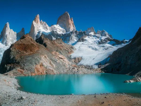 A tranquil turquoise lake with a backdrop of rugged snowy mountain peaks under a clear blue sky. Hire a travel agent to experience this serene landscape.