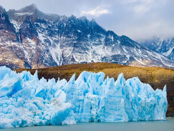 Hire a travel agent to visit the stunning scene of a jagged glacier in the foreground with snow-covered mountains in the background.