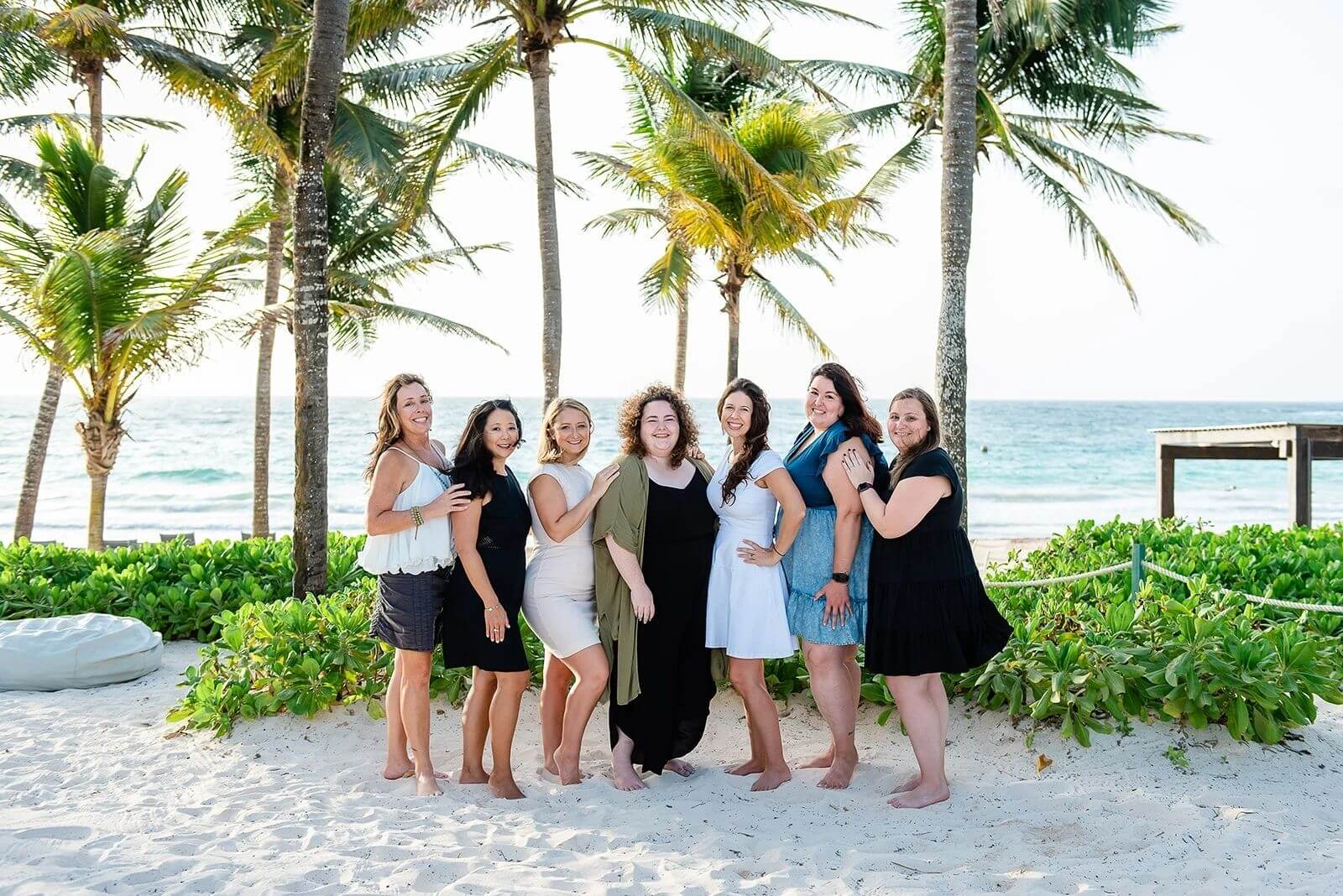 A group of women smiling together on a beach with palm trees in the background.