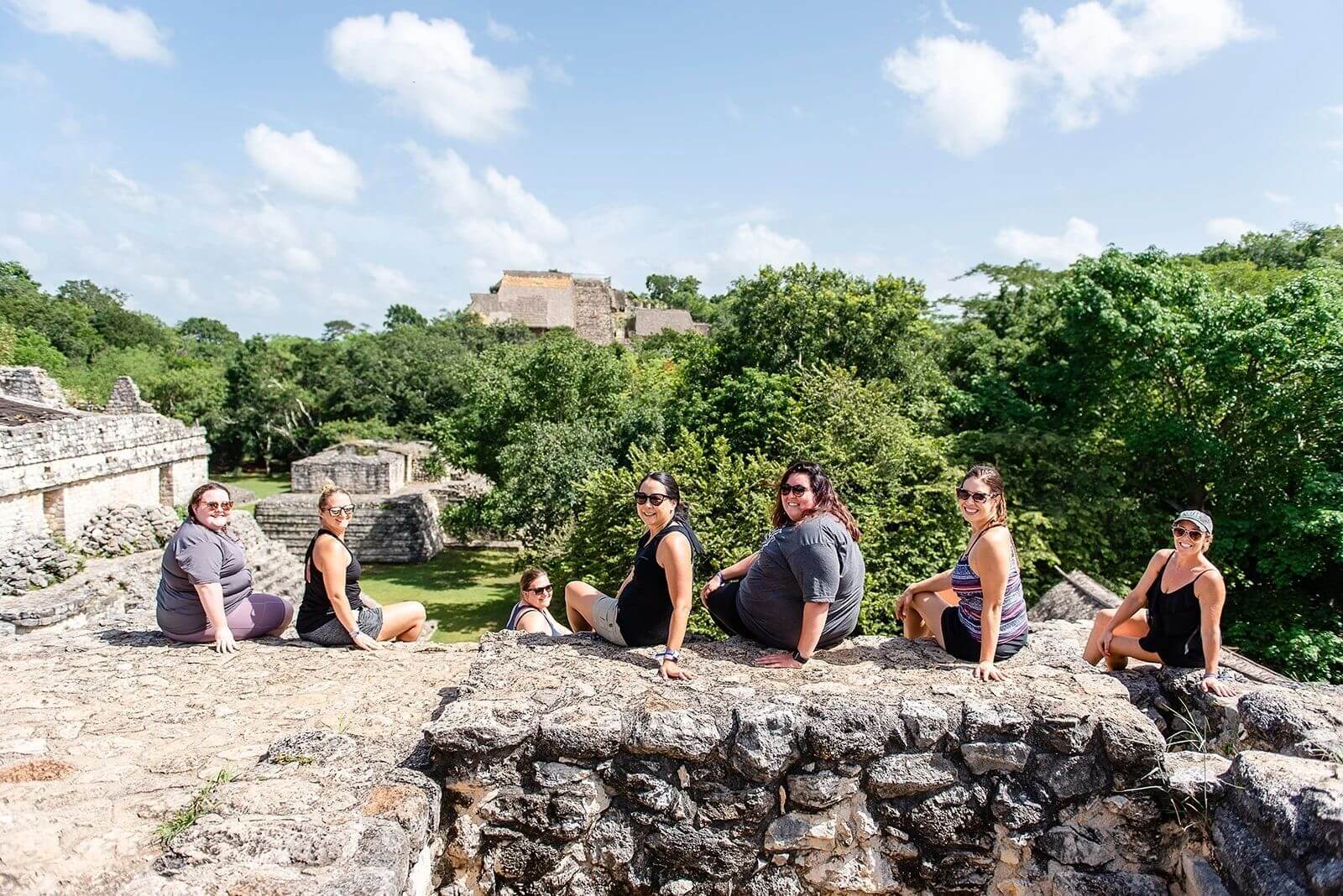 Group of tourists sitting on ancient ruins with a view of a mayan pyramid in the background.