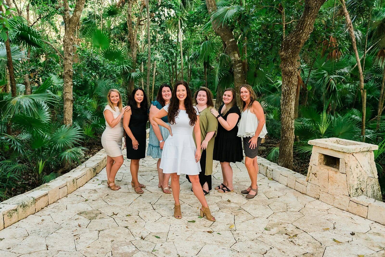 A group of women smiling and posing together on a stone pathway surrounded by lush greenery.