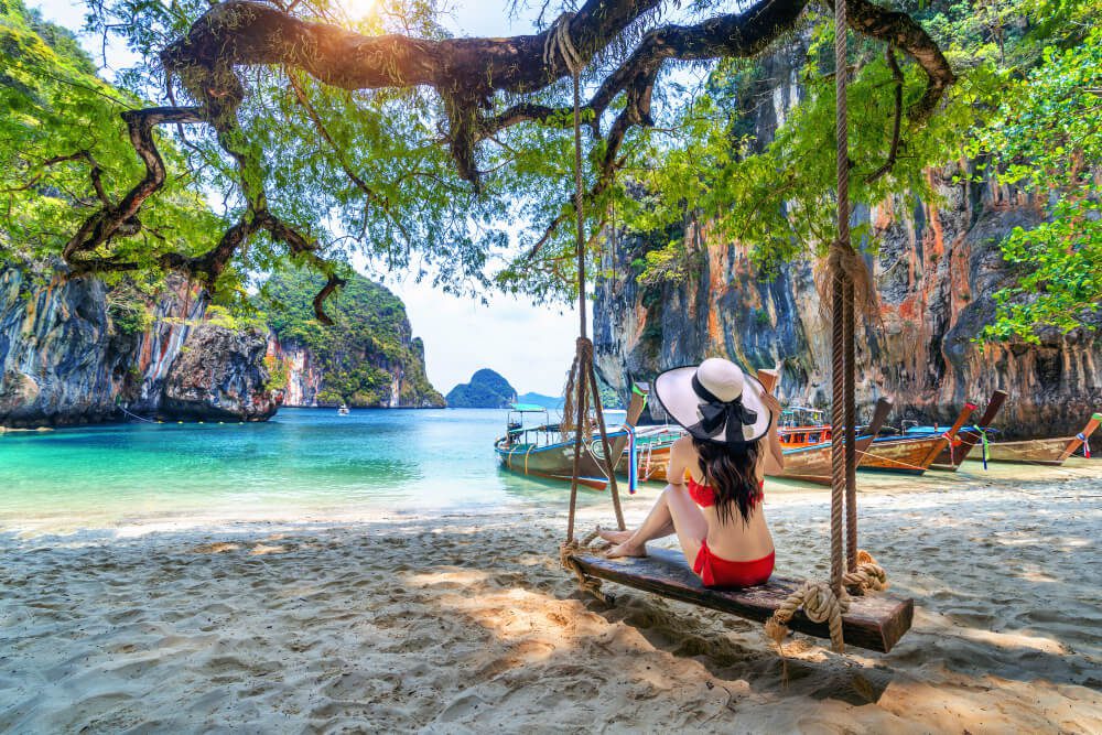 Woman in a sunhat sitting on a swing at a tropical beach with limestone cliffs and boats in the background, ready to become a travel agent.