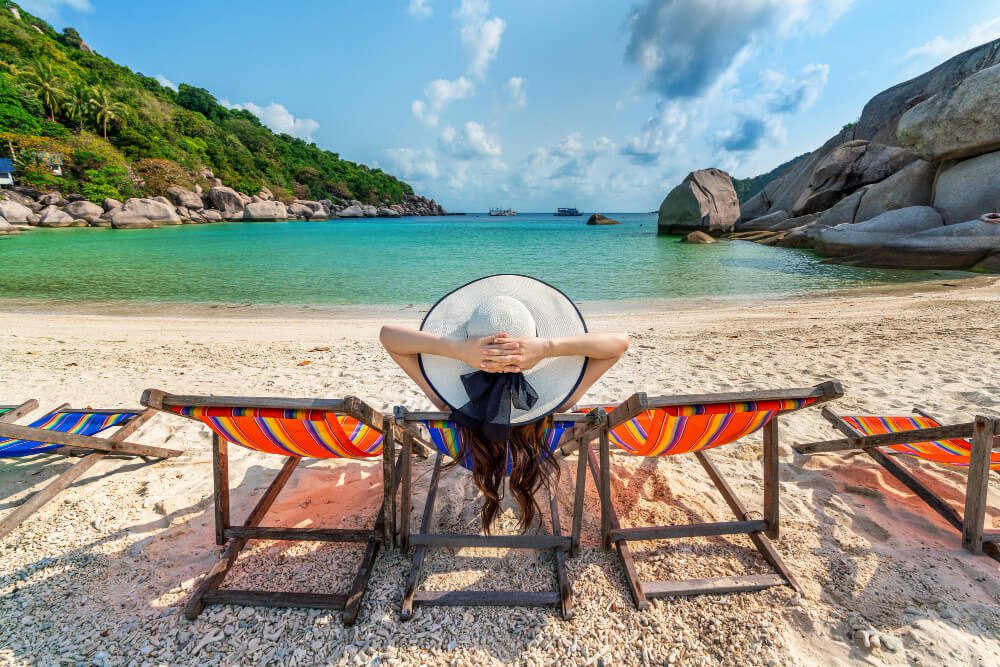 Person relaxing on a beach chair facing the sea with a wide-brimmed hat covering their head, dreaming of becoming a Travel Agent.