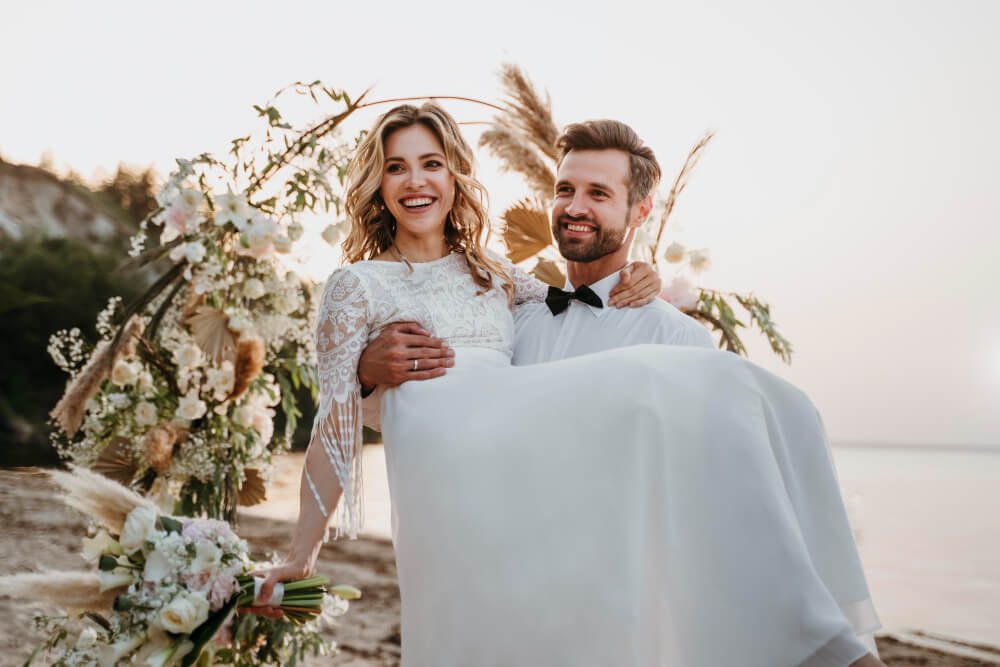 Groom carrying bride near a floral arrangement on the beach at sunset during their destination wedding.