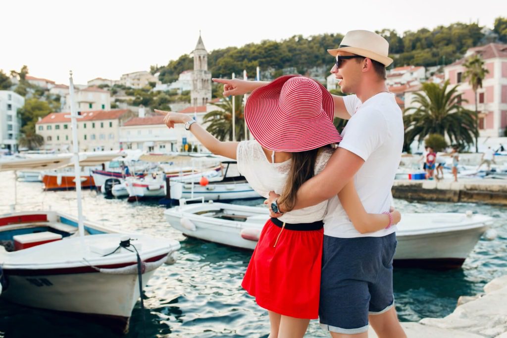 A couple in summer attire sightseeing at a harbor with boats and buildings in the background during their honeymoon.