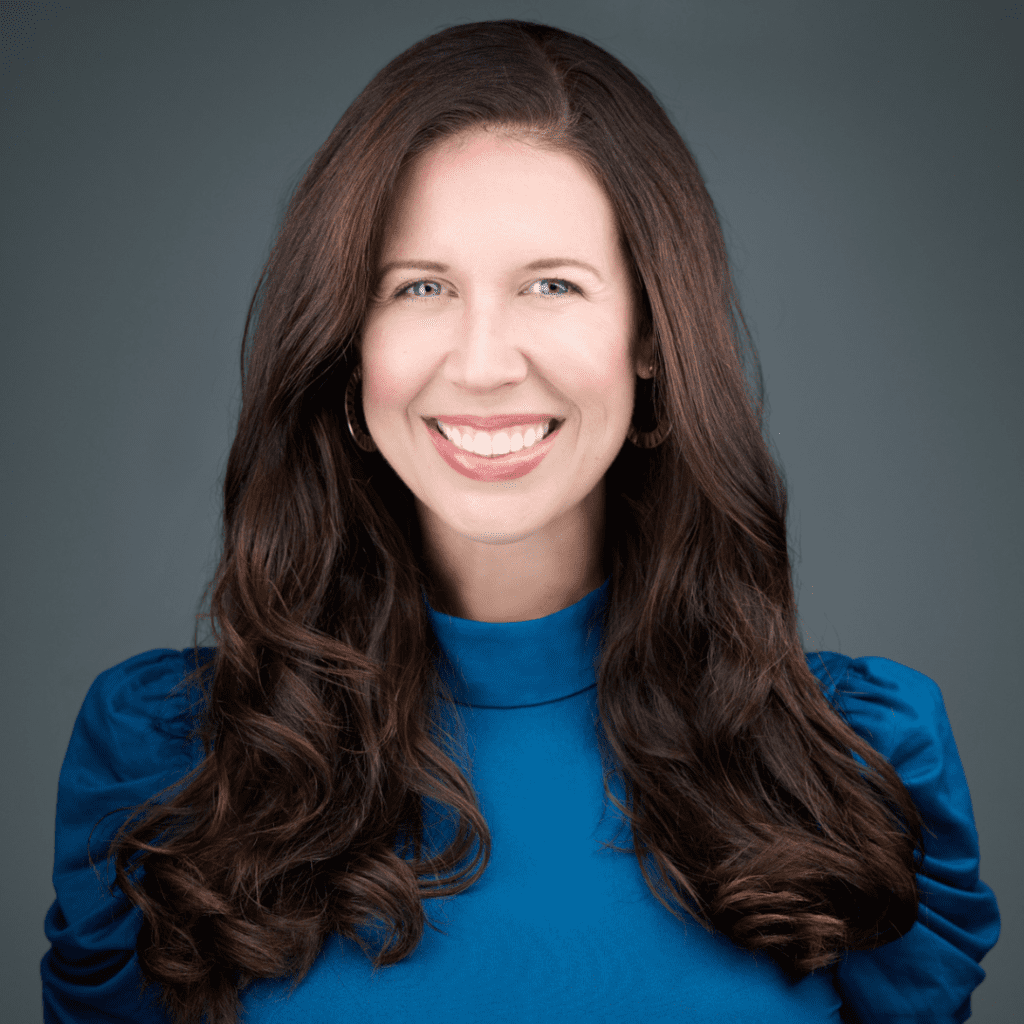 Professional woman with long curly brown hair wearing a blue top, smiling against a grey background.