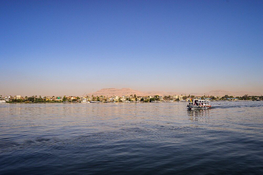 A boat navigating the nile river with a backdrop of a desert landscape and buildings on the riverbank.