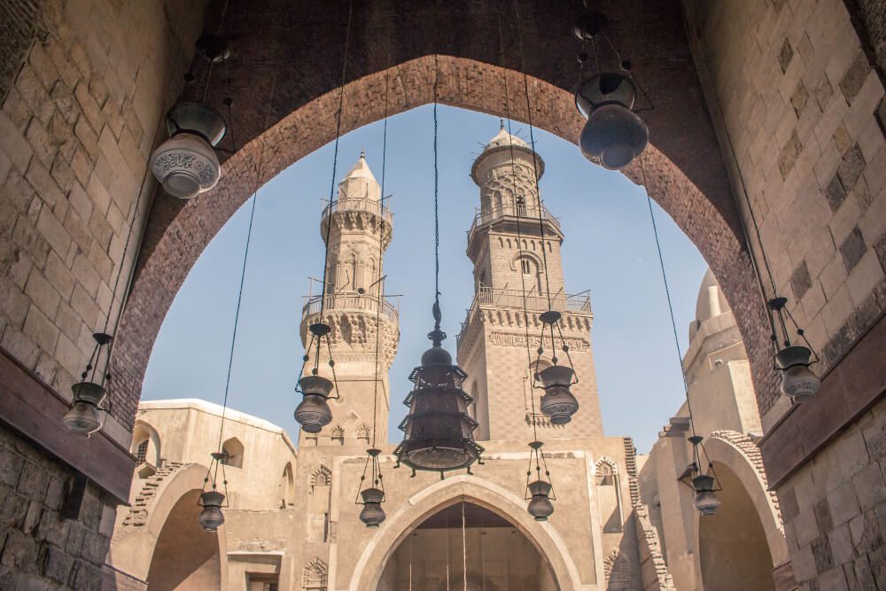 Arched gateway framing a view of traditional minarets and hanging lanterns against a clear sky.