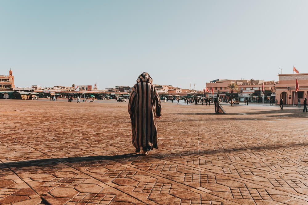 A person wearing traditional clothing walks across a spacious town square under a clear sky.