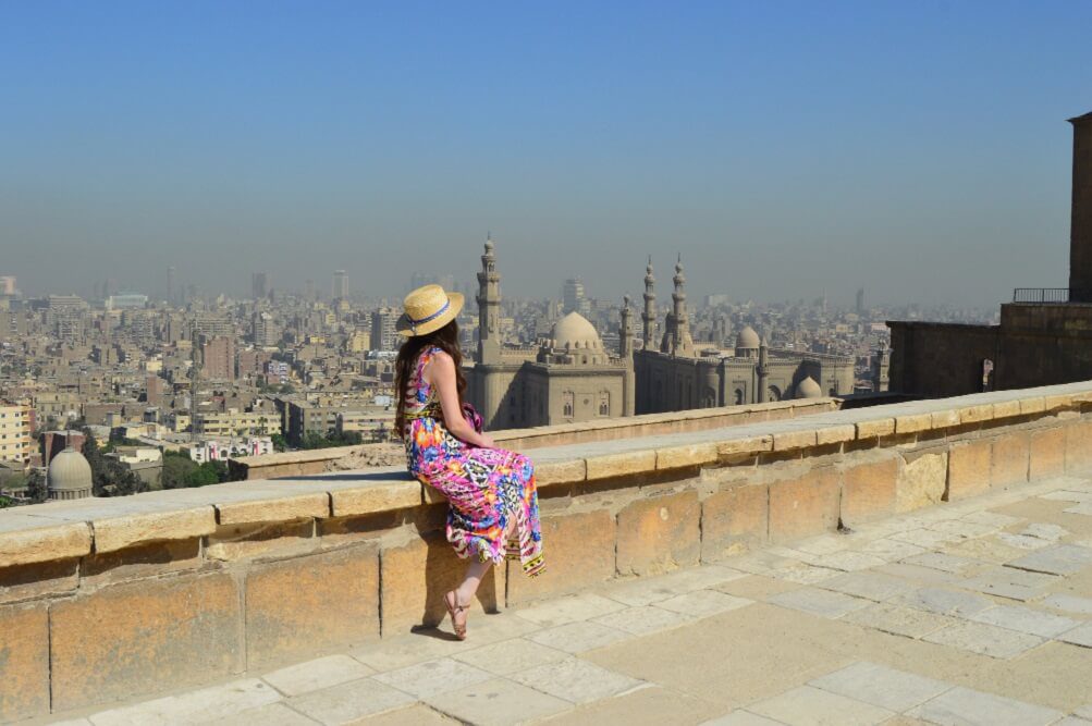 Woman wearing a summer dress and hat overlooking a historic cityscape from a high vantage point.