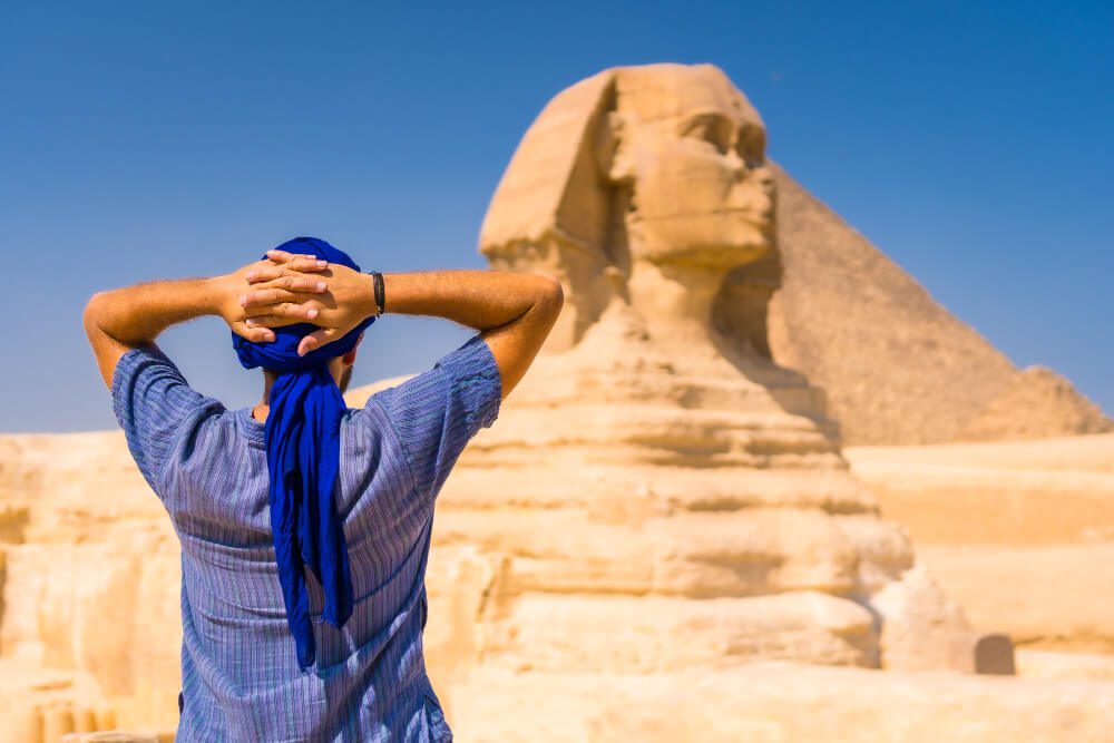 Tourist in a blue headscarf standing with hands behind head, gazing at the great sphinx of giza under a clear blue sky.