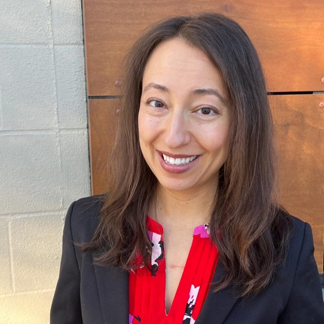 A smiling woman with long hair wearing a red blouse and black blazer.