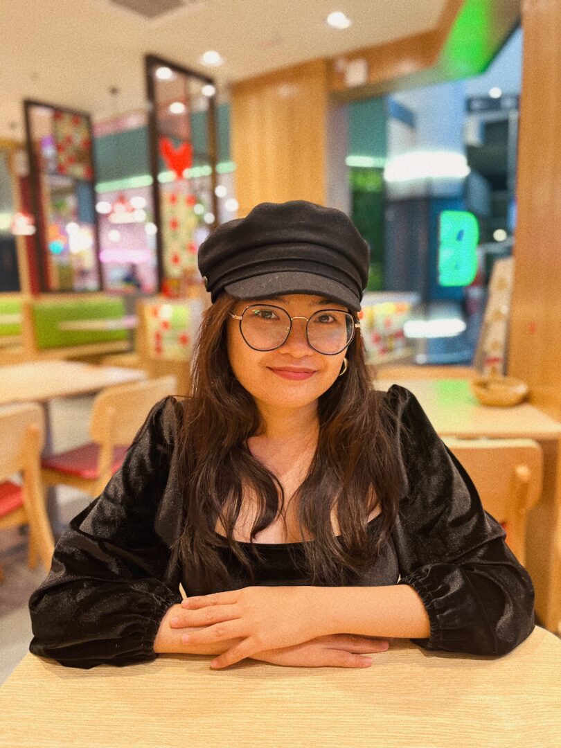 A smiling travel expert wearing a black cap and glasses seated at a table in a brightly lit food court.