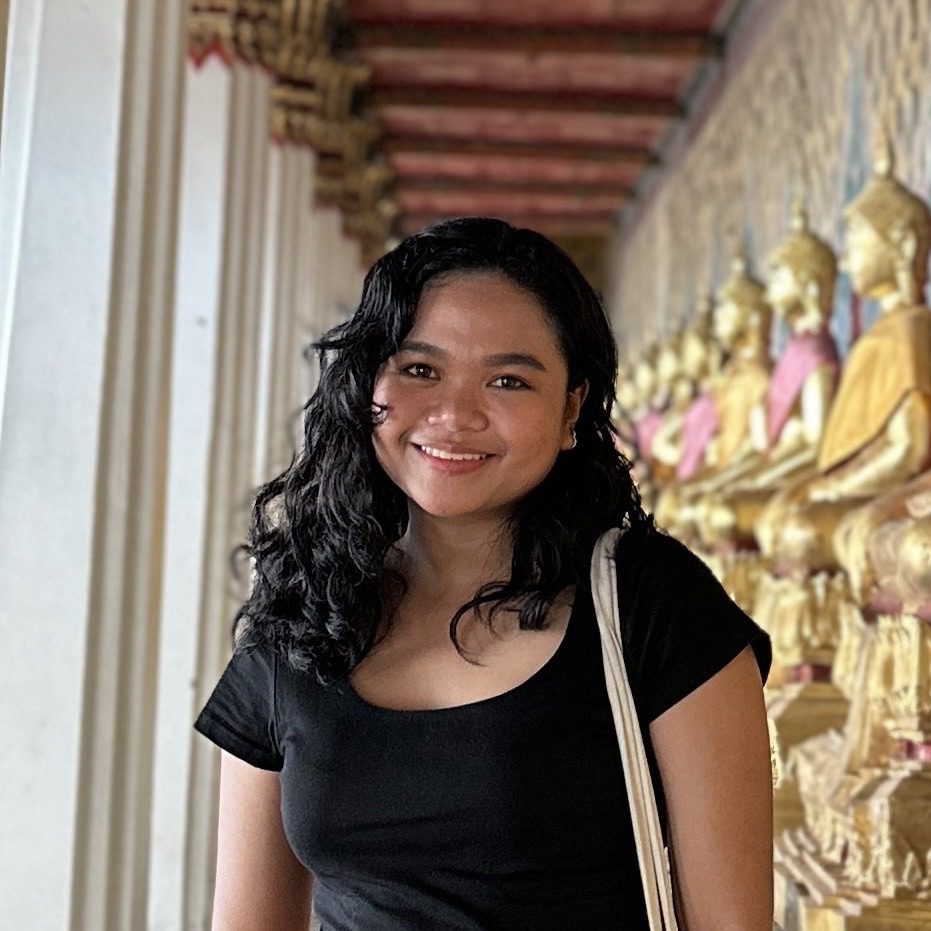 A smiling woman with curly hair, a travel expert, standing in a corridor with traditional sculptures and architectural details in the background.