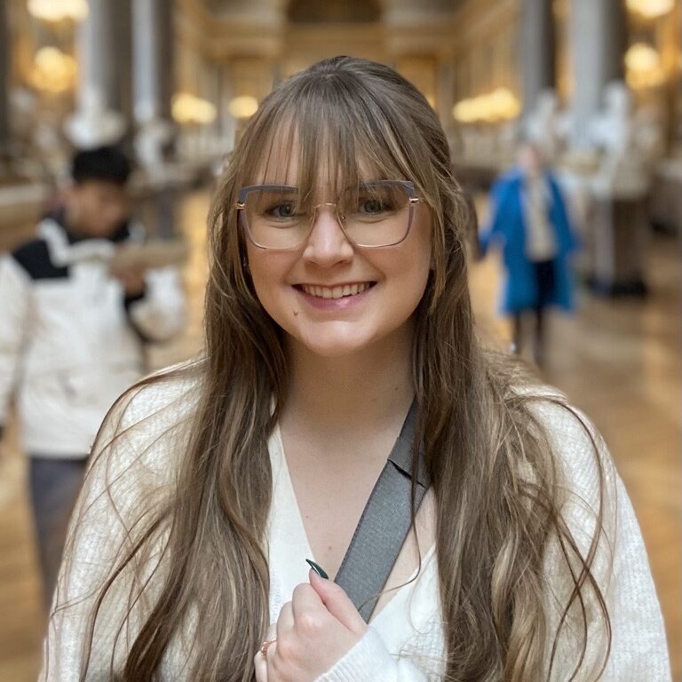 A smiling woman with glasses standing in an art gallery, consulted by travel experts.