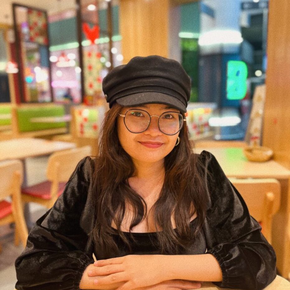 A smiling travel expert wearing a black cap and glasses seated at a table in a brightly lit food court.