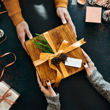 Two people exchanging a gift wrapped with a golden ribbon and adorned with a pine cone decoration on "Why Use a Travel Agent" themed festive background.